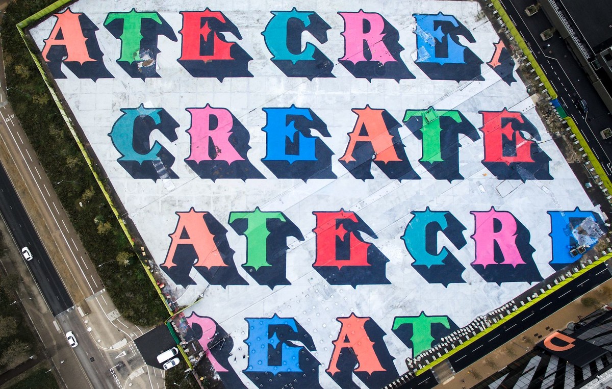 ベン・アイネ『CREATE』を空から見た写真。白い地面いっぱいに、水色やピンク、赤、緑、青の黒い影つきの文字でCREATEが何段にも繰り返され、脇の道路に車が小さく見える