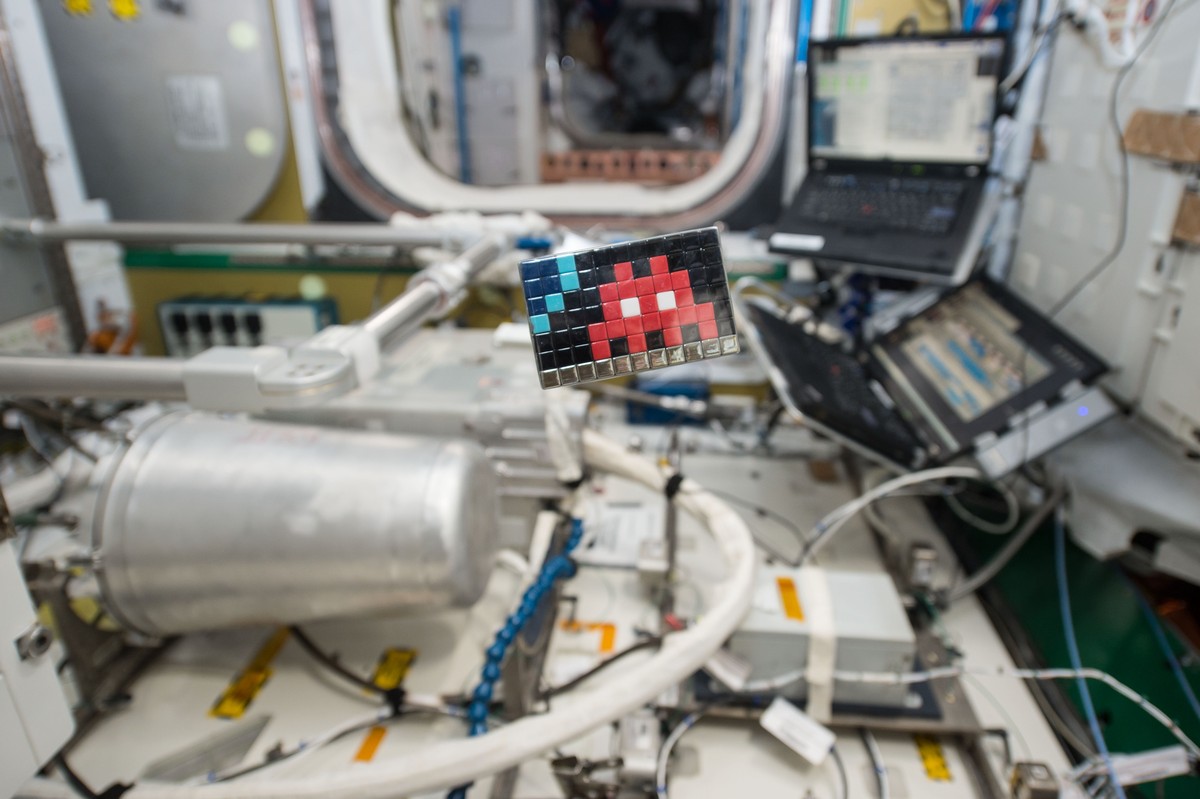 インベーダー『Space2』。ケーブルや機器、ノートパソコンが並ぶ国際宇宙ステーションの内部に、黒い地に赤いインベーダーを描いた小さなモザイクが設置されている写真