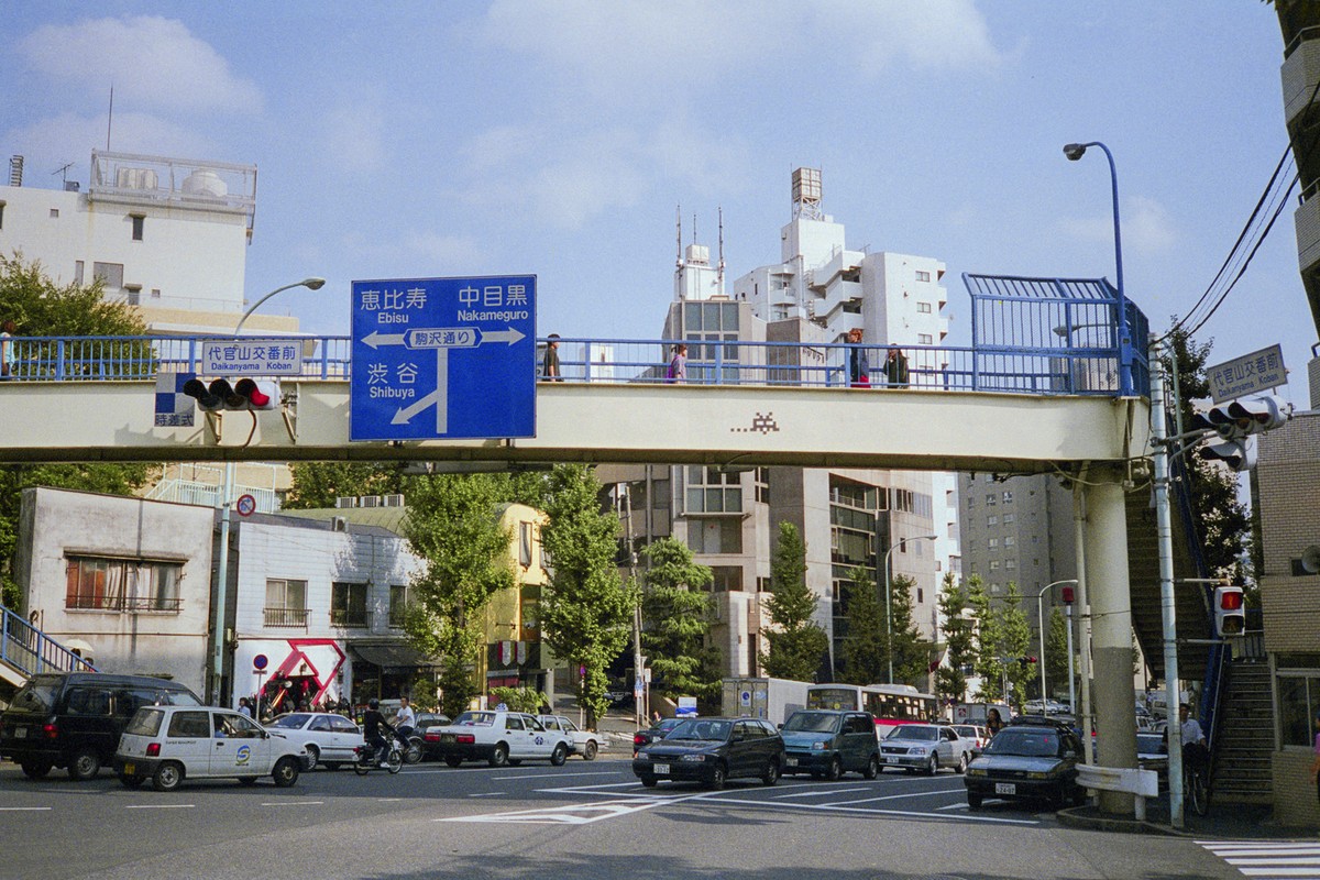 インベーダーの東京の作品。青い道路標識が掛かった歩道橋の側面の中央に小さなモザイクが貼られ、手前の道路を車が走る街の写真