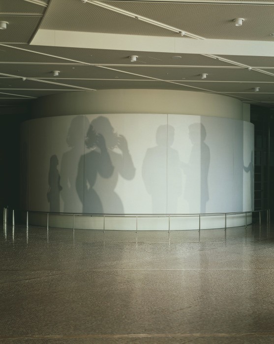 広い空間に据えられた円筒形の白い壁面に、人の姿の影だけが並ぶ壁画。左に電話をかけるような女性、右に向かい合う男性ふたりの影が見える。
