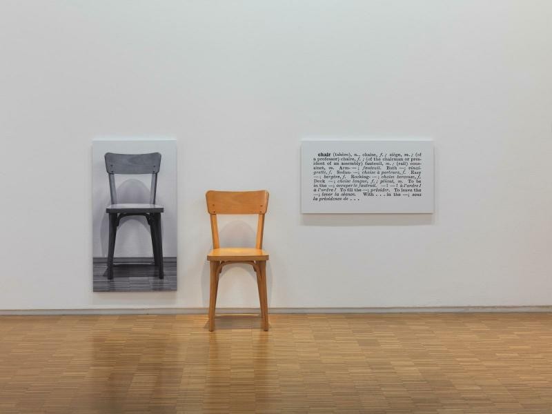 ジョセフ・コスース『One and Three Chairs』の展示風景。白い壁の左に椅子の白黒写真、その前の床に明るい色の木の椅子、右の壁に辞書の「chair」の項目を拡大した写真が並ぶ