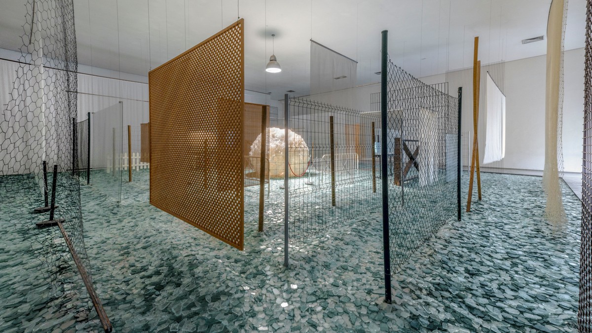 シルド・メイレレス『Através』の展示風景。割れたガラス片が敷き詰められた床の上に、木の格子の板、金網のフェンス、白い柵、薄い布などの仕切りが何重にも立ち、その奥の中央に丸めたセロハンの大きな白い球が見える