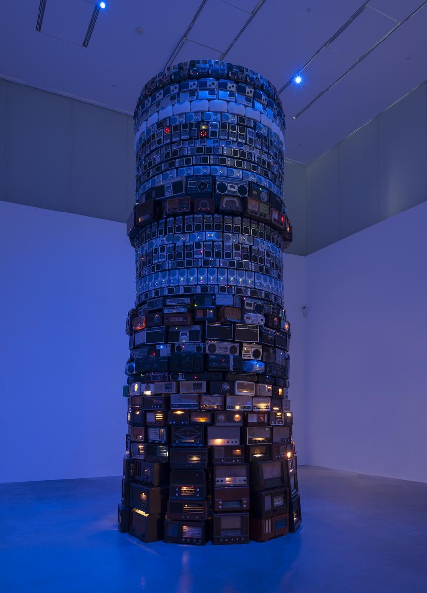 シルド・メイレレス『Babel』の展示風景。青い光に照らされた展示室の中央に、大小さまざまな古いラジオを何層にも円く積み上げた、天井近くまで届く塔が立ち、ラジオの小さな明かりが点々と光っている