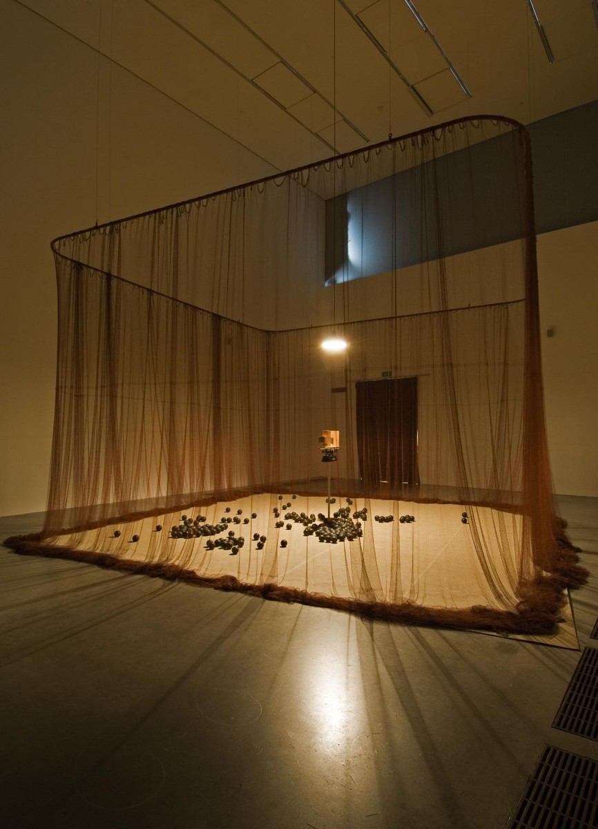 シルド・メイレレス『Eureka/Blindhotland』の展示風景。暗い展示室の中央に天井から下がる薄茶色の網が四角く囲いをつくり、その内側の床に黒いゴムのボールが散らばり、奥の中央に明かりに照らされた天秤が立つ