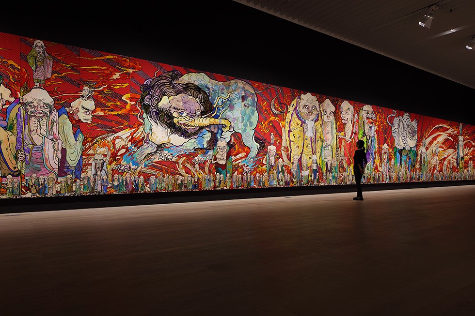 暗い展示室の壁一面に、赤い炎と多数の羅漢が極彩色で描かれた全長100メートルの巨大絵画『五百羅漢図』の一部。右に鑑賞者の人影