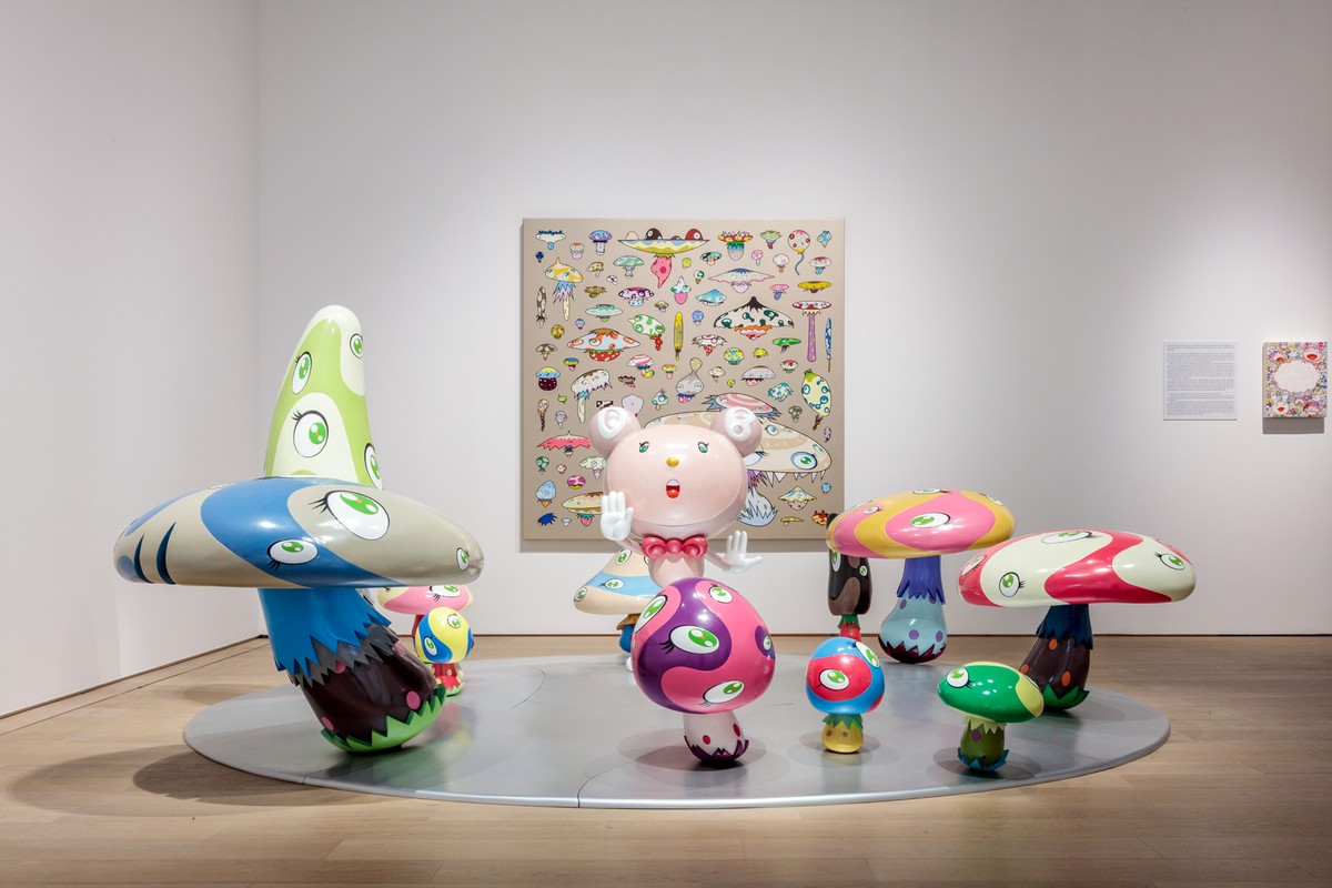 白い展示室の床に置かれた、目のついた色とりどりのキノコの彫刻群。中央に白いキャラクター、奥にキノコ柄を散りばめた大きな絵画が掛かる
