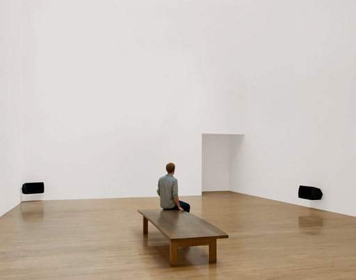 スーザン・フィリップス『Lowlands』のテート・ブリテンでの展示風景。白い展示室の壁ぎわに黒いスピーカーが2台あり、中央の木のベンチに来場者が1人座っている