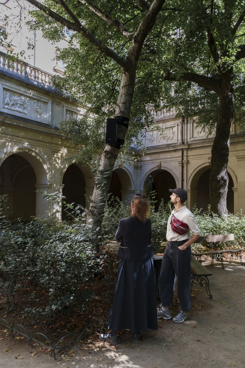 スーザン・フィリップス『We are the weavers』のリヨン美術館の庭園での展示風景。アーチの回廊に囲まれた庭の木の幹に黒いスピーカーがあり、2人が見上げている