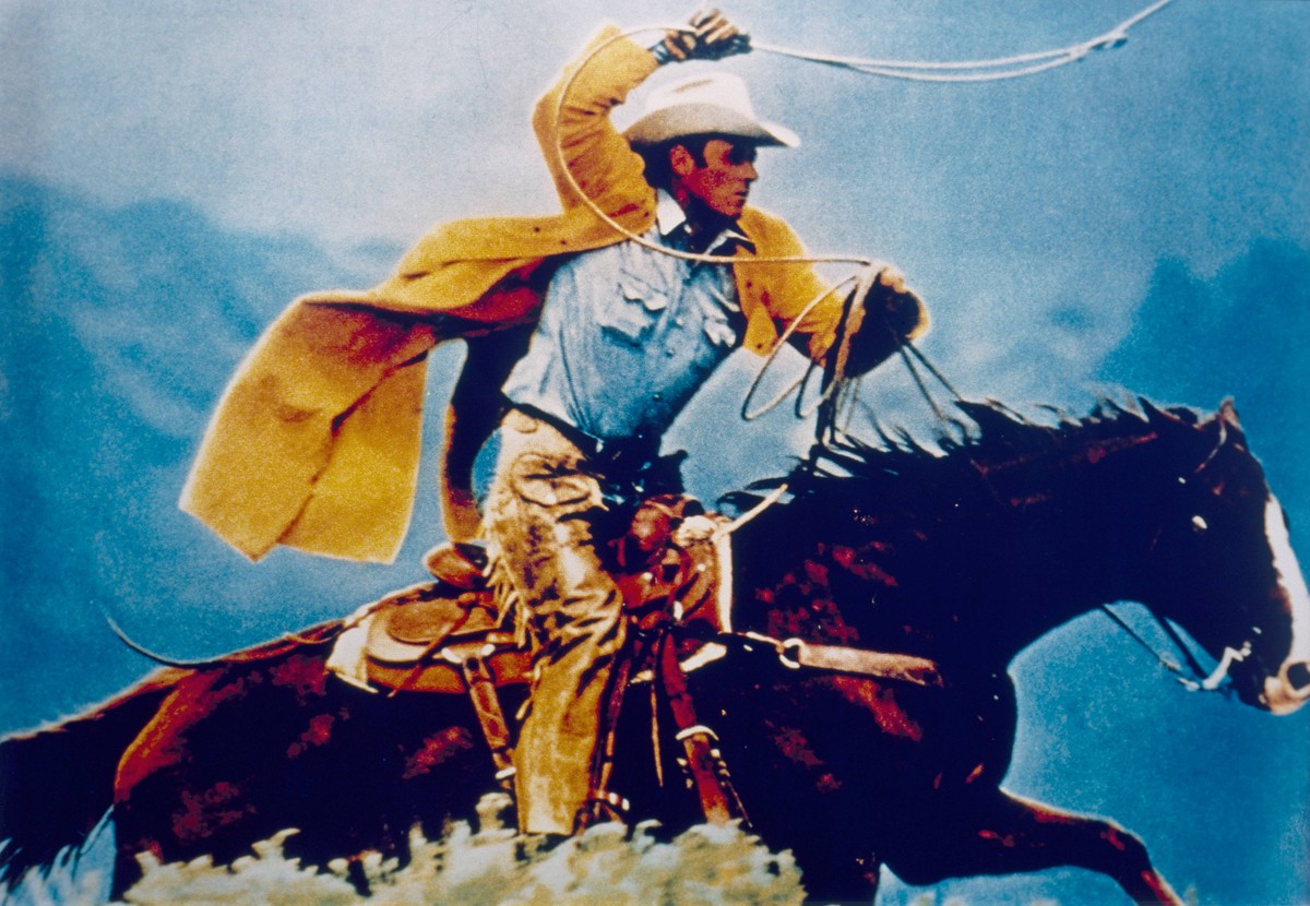 リチャード・プリンス『Untitled (Cowboy)』。青い空と山並みを背景に、白いカウボーイハットに黄色いコートのカウボーイが茶色の馬を走らせ、投げ縄を頭上で振り回すカラー写真