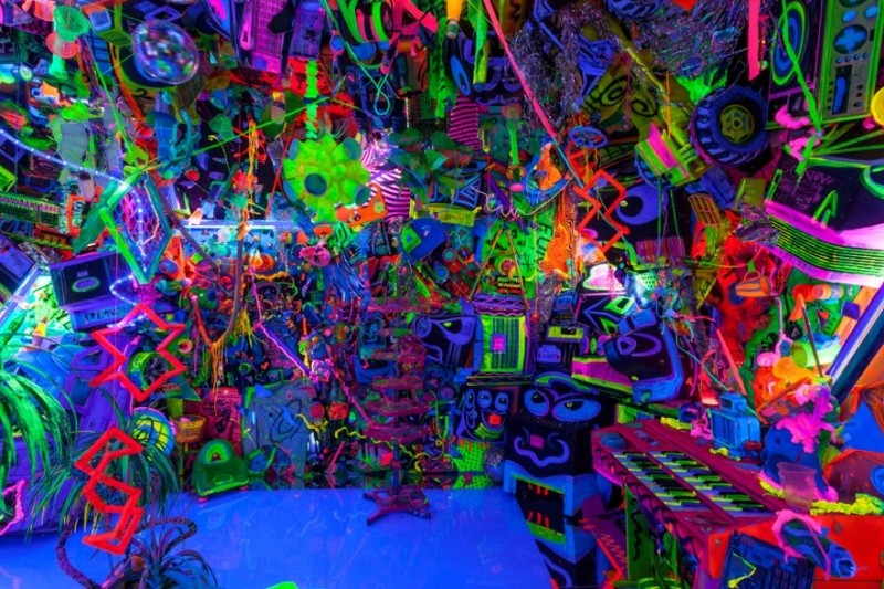 ケニー・シャーフ《Cosmic Cavern》第33作の内部。蛍光色の網やスピーカー、鍵盤楽器、おもちゃ、人形の顔が床から天井まで重なり、青紫のブラックライトの中で光っている部屋