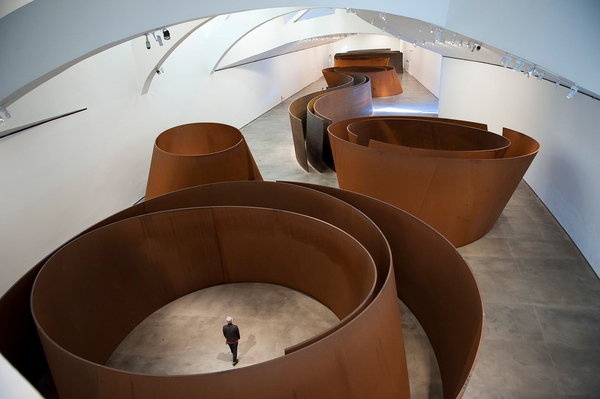 ビルバオ・グッゲンハイム美術館の展示室に並ぶ、赤茶色に錆びた巨大な鋼板の渦巻き。高い曲面の壁の中央に小さく人が立っている。