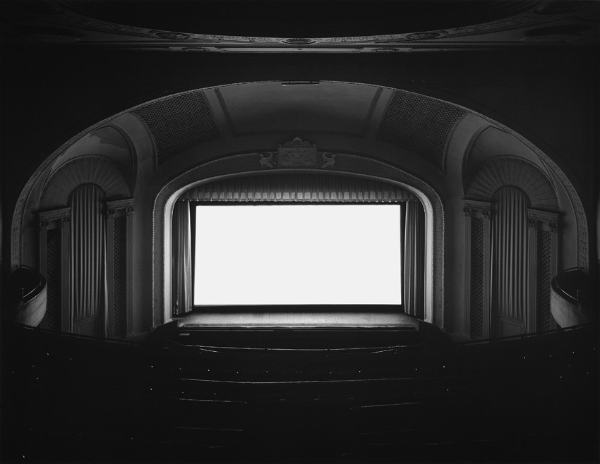 無人の古い映画館の客席から見たモノクロ写真。アーチ型の壁の奥で、上映された映画一本分の光がスクリーンを真っ白な長方形に輝かせている